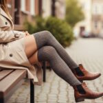 Młoda kobieta na ławce w beżowym płaszczu, z założonymi modnymi wysokimi skarpetami i skórzanymi botkami
